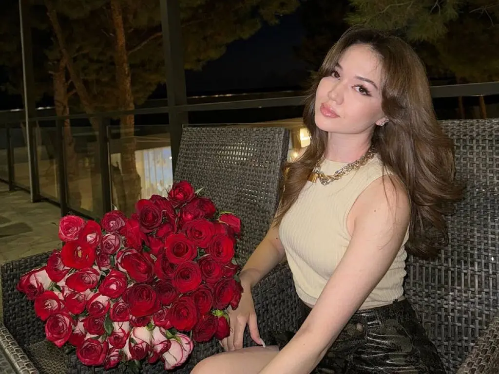 Young woman in a beige sleeveless top sitting on a patio chair next to a large bouquet of red roses at night.