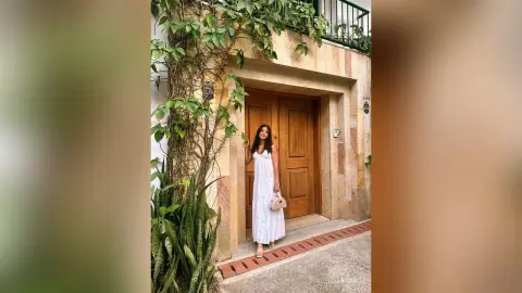 Elegant woman in a white summer dress standing by a rustic wooden door in a lush garden setting.