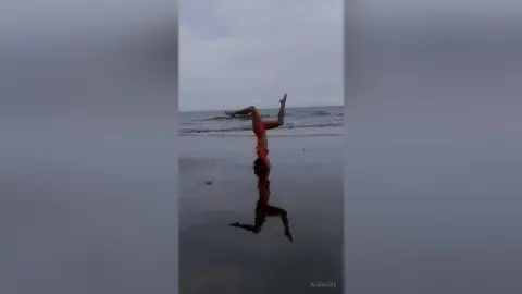 Athletic woman in a red bikini performing a handstand on a reflective beach at dusk.
