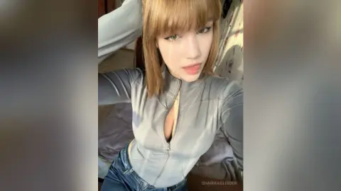 SharikaGlidden licks her lips while wearing a tight, long-sleeved grey zip-up top and blue jeans.