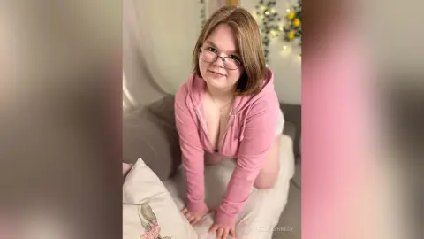 ManeelaKennedy wears glasses and a pink hoodie while kneeling on a white pillow in her bedroom.