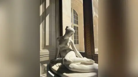 Classical white marble sculpture of a seated woman in a sunlit, elegant museum setting with soft shadows.