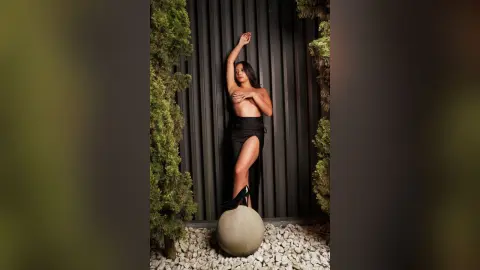 Elegant woman in a black slit dress posing against a dark slatted wall in a moody garden setting.