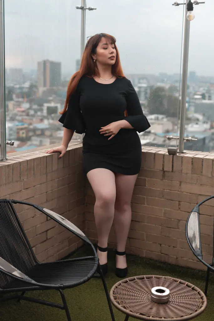 NiicolleEvanss with long brown hair wearing a black long-sleeve top and black shorts, sitting on a chair on a balcony overlooking a city.
