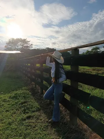CeleGarcia poses in a white shirt and blue jeans while leaning against a wooden fence under a bright sky.