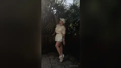 BiankaPerese stands outdoors against a backdrop of lush green tropical leaves, wearing a white short-sleeved top, white shorts, and a white hat.