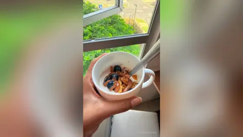 Hand holding a white cup of yogurt topped with blueberries and granola by a bright window overlooking green trees.