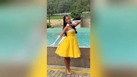 TefyRusso poses in a yellow strapless dress and long white gloves, standing by a swimming pool with a forest background.