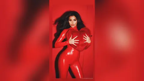 Glamorous woman in a red latex bodysuit holding a large plush heart against a vibrant red background.
