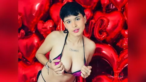 Young woman with short dark hair in black bikini posing against a backdrop of red heart balloons.