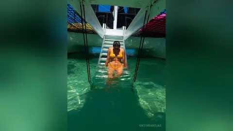 Young woman in an orange bikini relaxing in turquoise tropical water near a boat ladder on a sunny day.