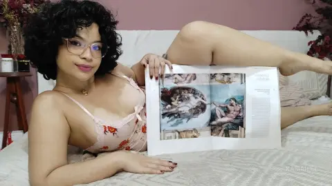 Curvy woman with glasses in a floral camisole relaxing on a white sofa while reading an art magazine.