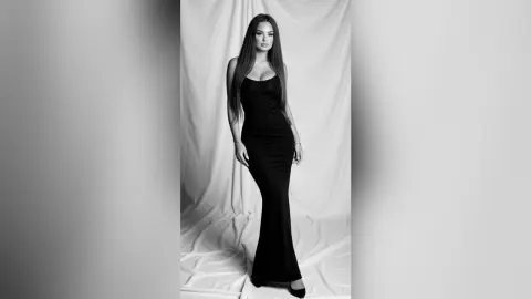 Elegant woman in a sleek black evening gown posing against a draped fabric backdrop in a moody black and white portrait.
