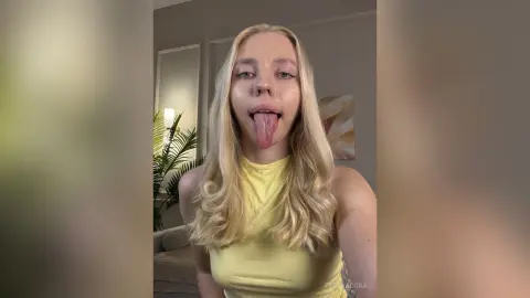 Blonde woman in a yellow halter top sticking her tongue out in a bright, casual indoor setting.
