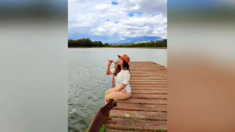 Woman in white blouse and sun hat relaxing on a wooden pier by a calm lake under a cloudy sky.