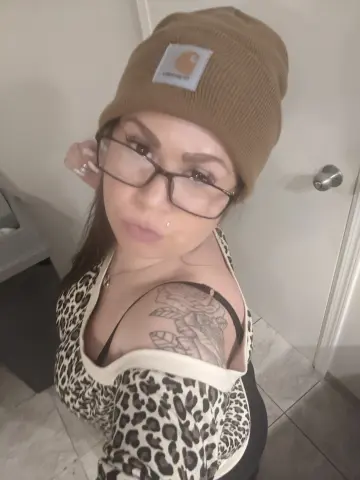Young woman with glasses and a tan Carhartt beanie wearing a leopard print top in a casual indoor setting.