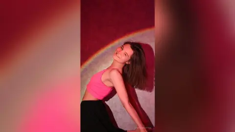 CaritaMattox smiles while posing in a pink crop top and black skirt against a colorful background.