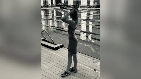 Young woman in a sleek bodysuit standing by an indoor swimming pool in a moody black and white aesthetic.