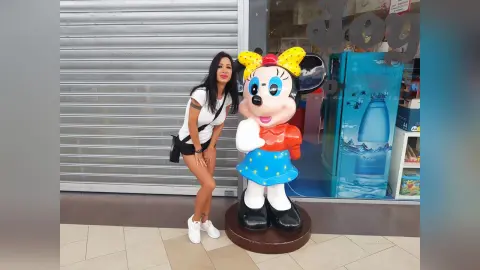 Woman in white t-shirt and black shorts posing next to a large Minnie Mouse statue in a bright storefront.