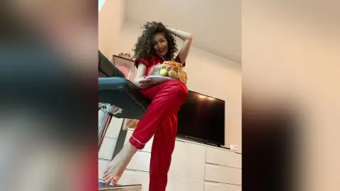 Smiling woman in red silk pajamas holding a gift basket in a bright modern living room.