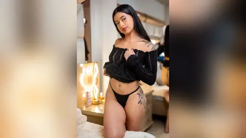 Young woman in black off-the-shoulder top and bikini bottoms posing confidently in a bright bedroom.