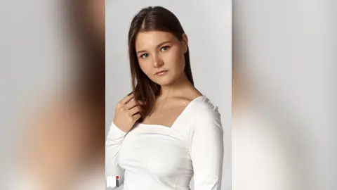 CaritaMattox poses in a white long-sleeve top with her brown hair down against a plain background.