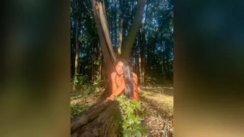 NicoleBella poses outdoors in a sunlit forest, wearing an orange top while leaning against a large tree trunk.