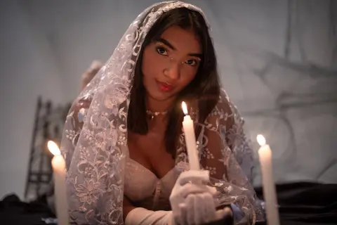 Ethereal woman in white lace veil and gloves posing with glowing candles in a moody, romantic setting.