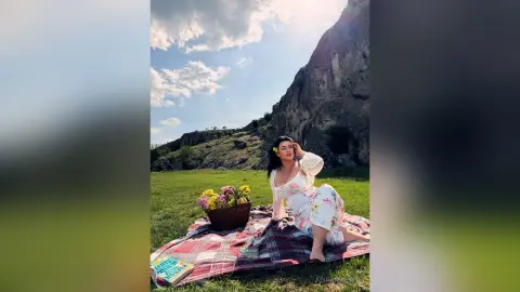 Woman in a floral sundress relaxing on a picnic blanket in a sunny meadow near rocky cliffs.