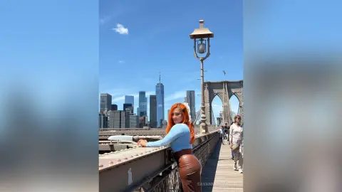 Redheaded woman in a blue sweater leaning on a railing with the New York City skyline and Brooklyn Bridge background.