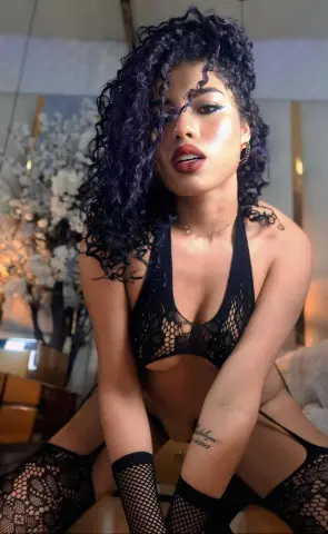 Curvy woman with curly dark hair wearing a black lace lingerie set in a bright, elegant bedroom setting.