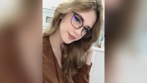 Smiling young woman with long blonde hair and black glasses wearing a brown shirt in a bright indoor setting.