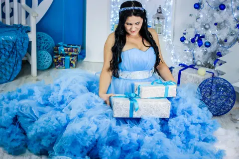 SweetEmmyGirl wearing a blue tulle dress and headband, sitting on the floor surrounded by silver gift boxes and a white Christmas tree.