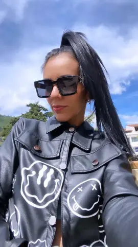 Woman in black leather smiley face jacket and sunglasses posing outdoors under a blue sky.