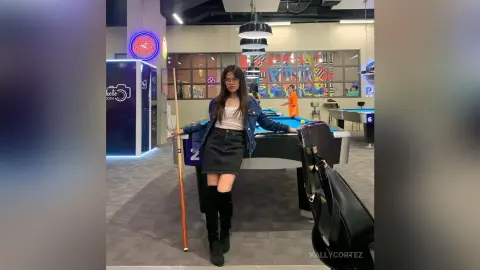 Young woman in denim jacket and black skirt standing by a pool table in a modern neon-lit game lounge.
