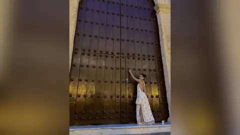Elegant woman in a white flowing gown standing before massive vintage wooden doors in a classic architectural setting.