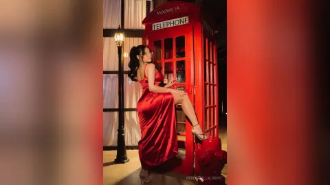 RebecaVillalobos poses in a red satin dress next to a red telephone booth, leaning back with one leg raised.