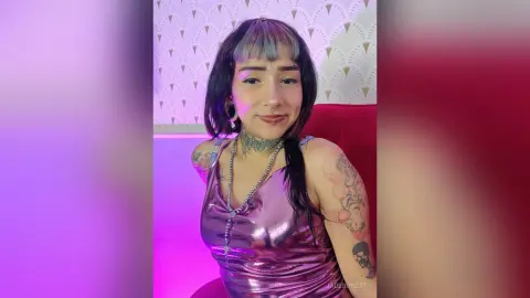 Smiling woman with dark bangs and neck tattoos wearing a metallic pink satin top in a soft purple light.