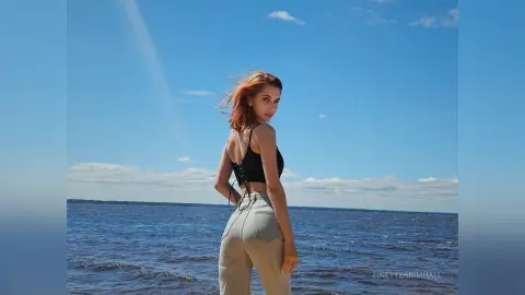 Redheaded woman in a black crop top and beige trousers standing by the blue ocean under a sunny sky.
