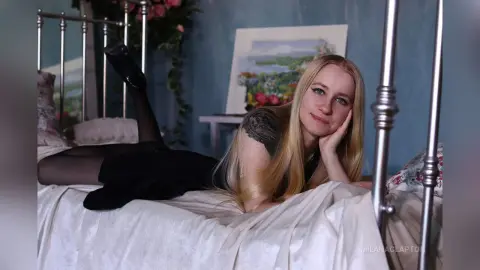 Blonde woman in a black dress relaxing on a white bed in a moody, blue-toned bedroom setting.
