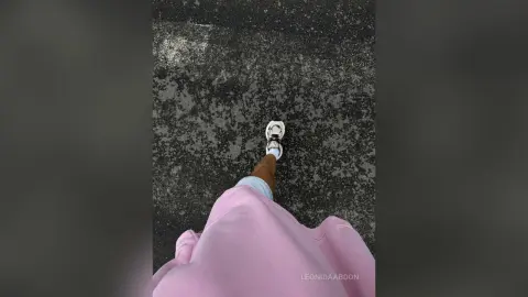 LeonidaAbdon wears a pink top and white sneakers while standing on a dark, textured pavement surface.
