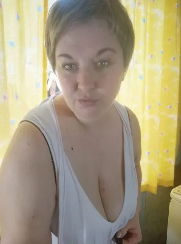 Juli_Bond wearing a white tank top with a deep neckline, showing her cleavage and short brown hair against a yellow curtain background.