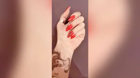 Close-up of a woman's hand with long red almond nails, a silver ring, and a detailed line art tattoo.