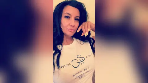MissyJolie poses in a white t-shirt with black text, looking at the camera with her hand near her face.