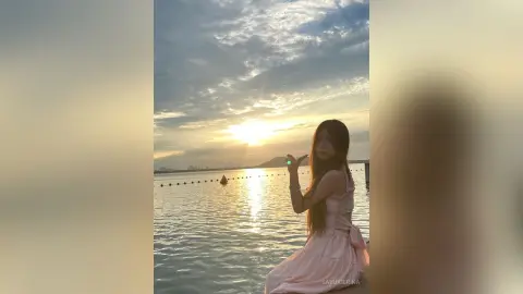 Young woman in a pink dress enjoying a peaceful sunset over the ocean at a scenic coastal lake.