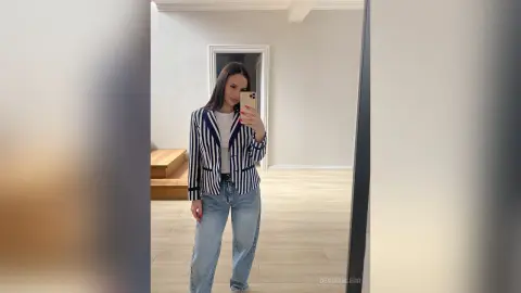 DeviaTelerr poses in a mirror wearing a blue and white striped blazer, a white top, and light blue jeans.