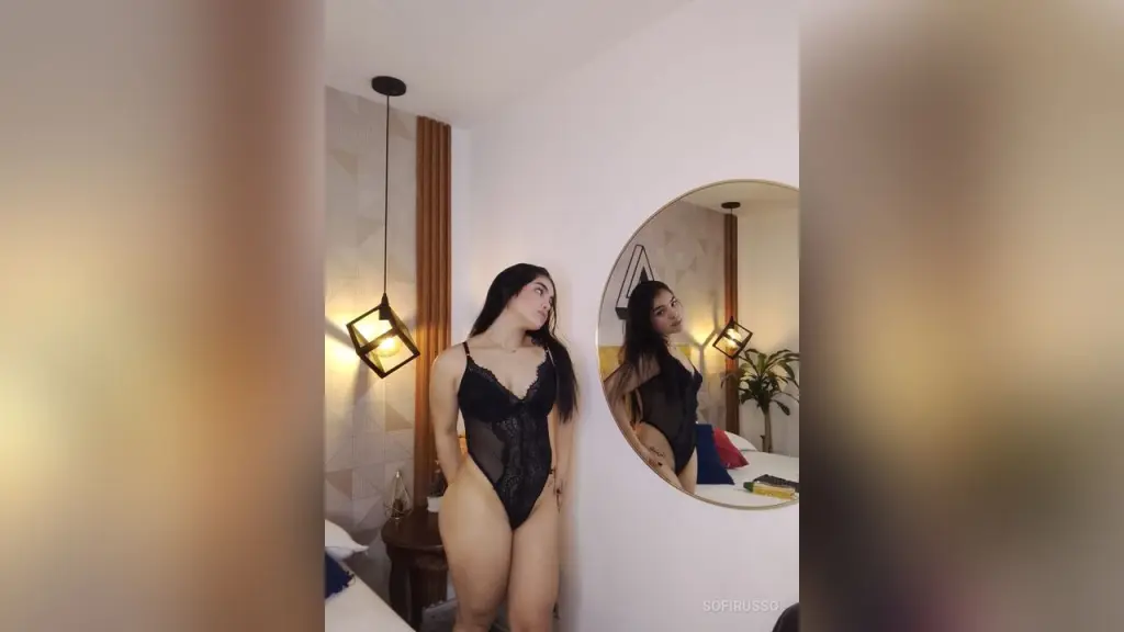 KimmieBloomie wears a white sailor top and dark skirt while sitting on a bed, showing off her cleavage and bare stomach.
