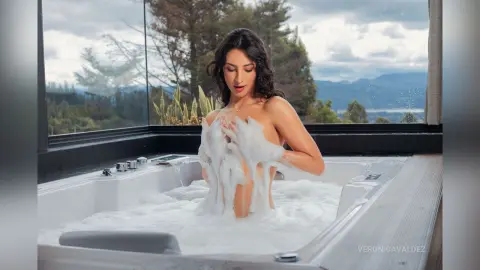 VeronicaValdez sits naked in a white bathtub filled with thick white soap bubbles, looking down with long dark hair.