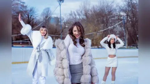 Three happy women in stylish white winter outfits laughing together at an outdoor ice skating rink.