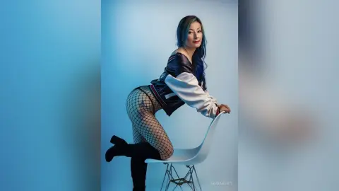 Stylish woman in black fishnet tights and a white top posing on a chair against a blue studio background.